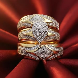May include: Three gold rings with diamond accents. The rings are stacked, showcasing a pear-shaped diamond centerpiece and diamond-studded bands. The rings are set against a red background.
