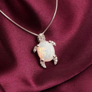 Op de afbeelding: Een zilveren ketting met een schildpadhanger. De hanger heeft een groot, ovaal, opalescent stenen lichaam, met diamantachtige accenten op de kop en vinnen. De ketting ligt op een dieprode stof.