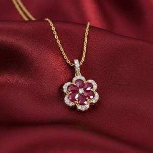 May include: A gold necklace with a flower pendant. The pendant features a cluster of red gemstones surrounded by a halo of small, clear stones. The necklace is displayed on a red satin background.