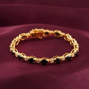 Puede incluir: Pulsera dorada con piedras negras ovaladas y pequeñas piedras claras. La pulsera está engarzada en una cadena dorada y tiene un cierre. La joya se exhibe sobre un fondo burdeos.