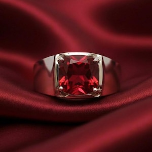 Może przedstawiać: Srebrny pierścionek z dużym, kwadratowym, ciemnoczerwonym kamieniem szlachetnym. Pierścionek ma grubą, wypolerowaną obrączkę, a kamień szlachetny jest osadzony czterema pazurami. Pierścionek jest prezentowany na czerwonej satynie.