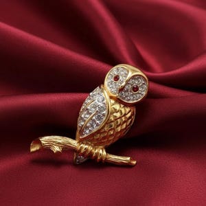 Pode incluir: Broche de cor dourada em forma de coruja com detalhes em cristais transparentes e vermelhos. A coruja está pousada num galho, com texturas detalhadas de penas e galhos. O broche está sobre um fundo de cetim vermelho escuro.