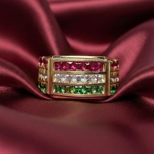 May include: A gold ring with three rows of square-cut gemstones. The top row features red gems, the middle row has white gems, and the bottom row is green. The ring is set against a deep red satin background.