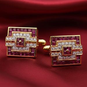 May include: A pair of gold cufflinks with a square design. Each cufflink features a central square of red gemstones surrounded by white gemstones and a gold frame. The cufflinks are set against a red background.
