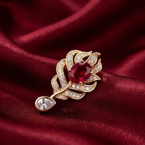 May include: A gold-toned feather-shaped brooch with a large, oval, red gemstone at its center. The brooch is encrusted with small, clear gemstones and features a pear-shaped clear gemstone at the bottom. The background is a deep red satin fabric.