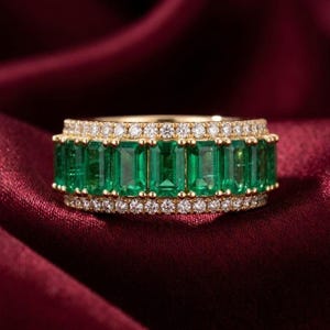 May include: A gold ring featuring a row of emerald-cut green gemstones, bordered by two rows of small, clear gemstones. The ring is set against a deep red background.