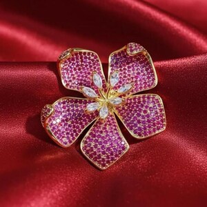 以下が含まれることがあります： 5枚の花びらを持つ金色の花のブローチ。各花びらは小さなピンク色の宝石で覆われています。中央には白いしずく型の石があります。ブローチは赤いサテンの背景に配置されています。