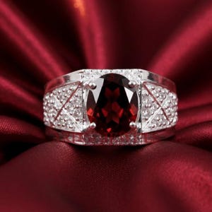 May include: A silver ring featuring a large, oval-cut, deep red gemstone. The band is embellished with smaller, clear stones. The ring is displayed on a rich, red satin surface.