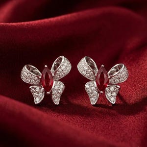 May include: A pair of silver bow-shaped earrings with a central, teardrop-shaped, deep red gemstone. The bows are encrusted with small, sparkling white stones, set against a rich, red background.