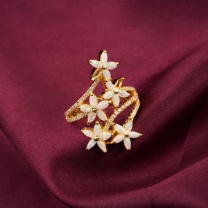 May include: A gold-toned ring featuring a floral design with five white flower accents. The ring has a curved band with small, sparkling embellishments. The ring is set against a deep burgundy fabric background.