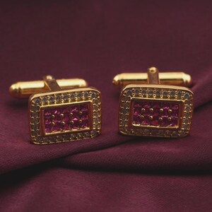 May include: A pair of gold-toned cufflinks with a rectangular design. Each cufflink features a central cluster of small, square, ruby-colored stones surrounded by a border of clear, sparkling stones. The cufflinks are set against a deep burgundy fabric.