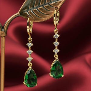 May include: Gold-coloured drop earrings featuring a green, teardrop-shaped gemstone and small, clear accent stones. The earrings are displayed against a red background, highlighting their elegant design and sparkle.