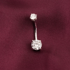 May include: A silver-colored belly button ring with two clear, round, sparkling gemstones. The curved barbell design features a larger stone at the bottom and a smaller one at the top, set against a deep burgundy background.