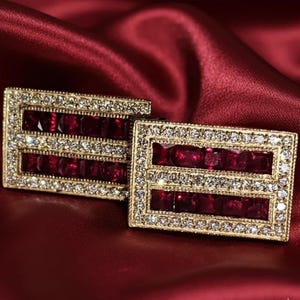 Może przedstawiać: Para prostokątnych, złotych spinek do mankietów z podwójnym rzędem prostokątnych, głęboko czerwonych kamieni szlachetnych. Krawędzie są inkrustowane małymi, przezroczystymi kryształami. Spinki do mankietów spoczywają na głęboko czerwonej satynowej powierzchni.