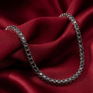 May include: A silver-toned necklace with a row of black gemstones. The necklace is displayed on a deep red satin fabric, creating a contrast between the shiny metal and the dark gemstones.