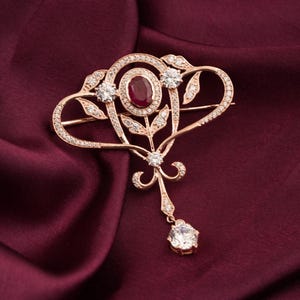 May include: A gold-coloured brooch with an oval ruby centre, encircled by clear crystals and intricate leaf patterns. The brooch has a dangling, round, clear crystal at the base. Set against a deep burgundy fabric.