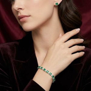 May include: A bracelet and matching earrings featuring square-cut emeralds and small, round, clear gemstones. The bracelet is worn on a wrist, and the earrings are studs. The jewelry is set in a silver or white metal.