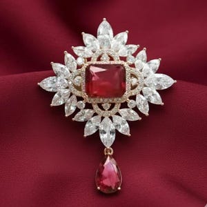 May include: A gold-toned brooch with a large, square, red gemstone at its center, surrounded by clear, petal-shaped crystals. A teardrop-shaped red gemstone hangs below. The brooch is set against a deep red fabric.