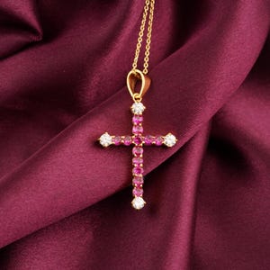 Pode incluir: Um colar com um pingente de cruz dourada. A cruz é adornada com pedras preciosas rosa e transparentes. O colar é exibido em um tecido de cetim vermelho escuro.