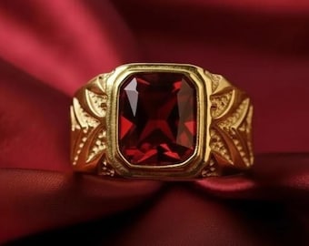 May include: A gold-colored ring with a large, rectangular, deep red gemstone. The ring has an ornate design on the sides and a polished finish, set against a red background. The gemstone is approximately 0.75 inches in length.