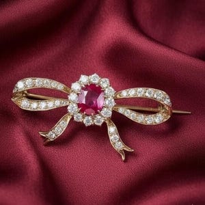 May include: A gold-toned bow brooch with a central oval ruby surrounded by a halo of diamonds. The bow's loops and tails are encrusted with smaller diamonds, set against a deep red satin background. A classic and elegant jewelry piece.