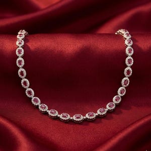 May include: A necklace featuring oval-cut red gemstones surrounded by small, clear stones. The necklace is set on a red satin background, creating a luxurious and elegant display. The gemstones are evenly spaced along the necklace.