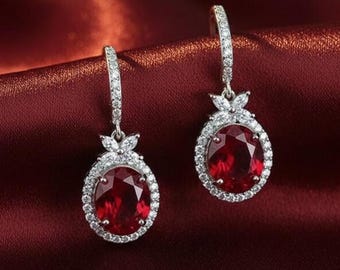 May include: A pair of silver-toned drop earrings with oval, deep red gemstones. Each earring is surrounded by a halo of small, clear stones and features a decorative floral design at the top. The earrings are displayed against a rich, red background.