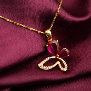 May include: A gold necklace featuring a butterfly-shaped pendant. The upper wings are set with two pear-cut deep red gemstones, while the lower wings are outlined with small, clear, sparkling stones set into the gold metal frame.