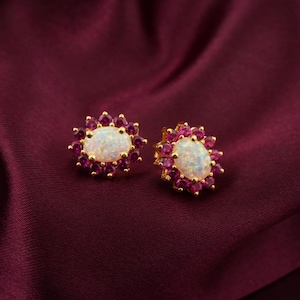 May include: A pair of gold-toned stud earrings. Each earring features a central oval-shaped opal surrounded by small, round, deep pink gemstones. The earrings are set against a dark red background.