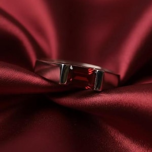 Puede incluir: Un anillo plateado con una gema rectangular de color rojo intenso engastada en una banda pulida. El anillo se exhibe sobre una rica tela de color burdeos, creando una estética lujosa. La gema mide aproximadamente 1,3 cm de largo.