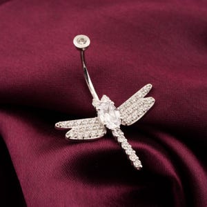 Puede incluir: Un piercing para el ombligo plateado con forma de libélula. La libélula tiene una piedra central grande, de forma ovalada, y piedras más pequeñas en las alas y el cuerpo. La piedra superior es redonda.