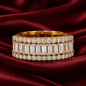 May include: A wide gold band ring featuring a central row of vertical baguette-cut clear stones flanked by two outer rows of small round brilliant-cut clear stones. The metal has a polished yellow gold finish throughout the entire ring structure.