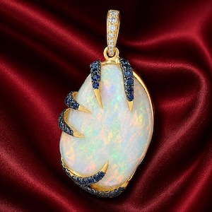 Puede incluir: Colgante de oro con una gran ópalo iridiscente. El ópalo está engarzado en un diseño de garra con piedras preciosas azules y detalles dorados. La fianza del colgante está adornada con pequeñas gemas claras.