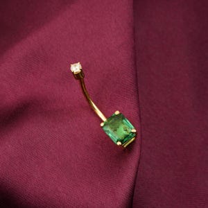 May include: A gold belly button ring with a curved design. It features a rectangular emerald-cut green gemstone and a small, round, clear gemstone. The jewellery is set against a deep burgundy fabric.