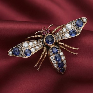 Puede incluir: Broche dorada con forma de abeja, con detalles intrincados. Las alas están adornadas con zafiros azules y gemas claras, con un gran zafiro en el centro del cuerpo. Dos pequeñas gemas rojas están en la cabeza.