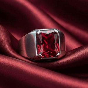May include: A silver ring with a large, rectangular, faceted, deep red gemstone. The ring is set on a rich, red satin fabric, creating a luxurious and elegant presentation. The gemstone's facets catch the light, displaying its brilliance.