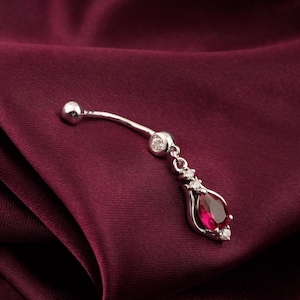 May include: Silver belly button ring with a teardrop-shaped, deep red gemstone. The pendant is accented with small, clear crystals. The curved barbell has a small silver ball at the top. The jewelry is displayed on a burgundy fabric.