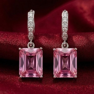 Puede incluir: Un par de pendientes plateados con gemas rosas rectangulares. Los pendientes tienen un diseño articulado con una fila de pequeñas piedras transparentes. El fondo es de tela roja oscura.