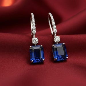 May include: A pair of elegant drop earrings featuring large, rectangular, deep blue gemstones. Each earring has a smaller, round, clear gemstone above the blue stone, and a row of smaller clear stones along the curved top. The earrings are set against a red background.