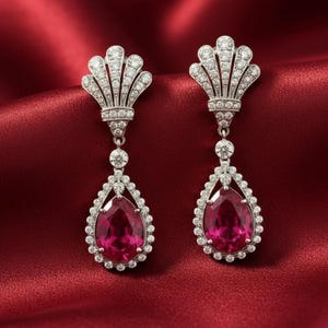 May include: A pair of elegant drop earrings featuring a fan-shaped top encrusted with small, sparkling stones. Each earring has a teardrop-shaped, ruby-colored center stone, surrounded by a halo of smaller, clear stones. The earrings are set against a red background.