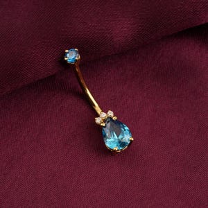 May include: A gold-coloured belly button ring with a curved barbell design. It features a teardrop-shaped, light blue gemstone at the bottom, accented with small clear stones. A smaller, round blue stone sits at the top.
