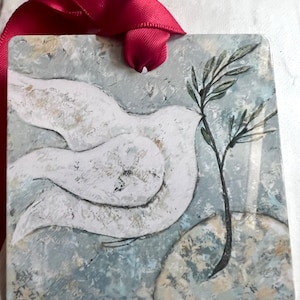 Könnte beinhalten: Ein weißer Keramik-Anhänger mit einer Taube und einem Olivenzweig. Die Taube fliegt mit ausgebreiteten Flügeln. Der Olivenzweig ist grün und hat Blätter. Der Hintergrund ist hellblau und grau.