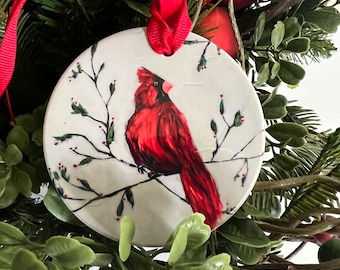 May include: A white ceramic ornament with a red cardinal painted on it. The ornament is hanging from a red ribbon and is surrounded by green foliage.