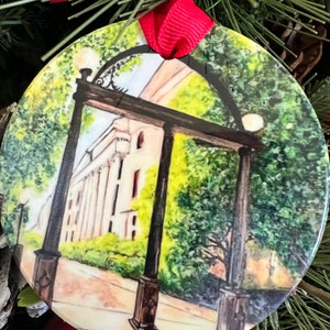 May include: A round Christmas ornament with a watercolor painting of a university archway and building. The ornament has a red ribbon for hanging and is surrounded by pine needles and a red ornament.