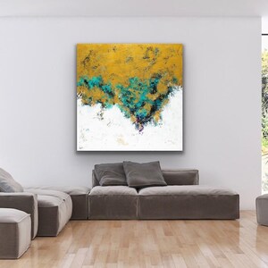 May include: Abstract art piece with a gold and teal textured design over a white background. The artwork is displayed in a modern living room setting, above a gray sectional sofa. The room has large windows.