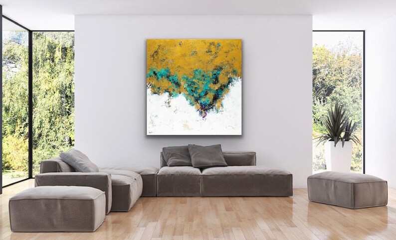 May include: Abstract art piece with a gold and teal textured design over a white background. The artwork is displayed in a modern living room setting, above a gray sectional sofa. The room has large windows.