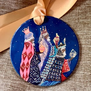 Puede incluir: Adorno navideño redondo y azul con tres reyes magos con ricas vestiduras, cada uno sosteniendo un regalo. El adorno tiene una cinta dorada para colgar. Los reyes están sobre un fondo de noche estrellada.