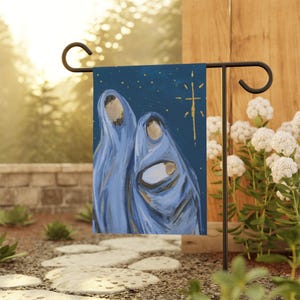 May include: A garden flag featuring an artistic depiction of the Holy Family. The painting shows Mary, Joseph, and baby Jesus in blue robes against a dark blue background with gold stars and a gold star of Bethlehem.