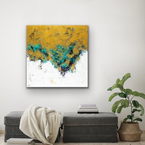 May include: Abstract art painting with a gold, teal, and white color palette. The artwork features textured brushstrokes and a layered composition. The painting is displayed on a white wall above two gray ottomans, a blanket, and a plant in a woven basket.
