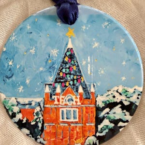 Pode incluir: Um ornamento redondo de cerâmica com uma cena pintada à mão de um edifício de tijolos com a palavra TECH em letras brancas. O edifício tem um telhado escuro com luzes coloridas e uma estrela amarela, num céu azul claro com flocos de neve.